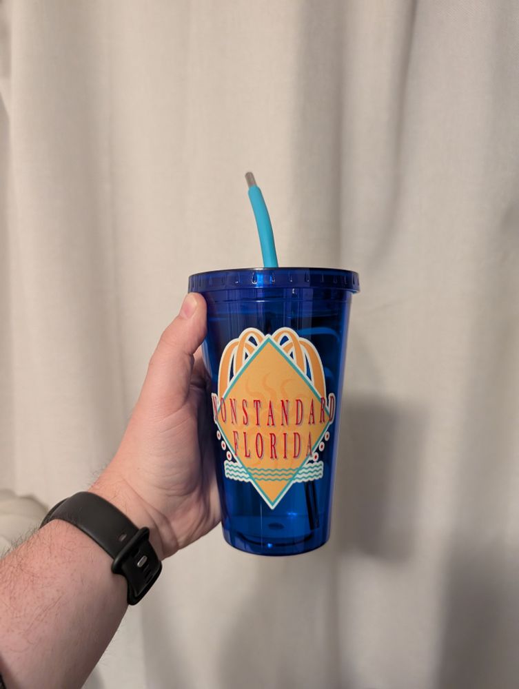 A man's hand holding a blue reusable cup with the words "Nonstandard Florida" on it. 
