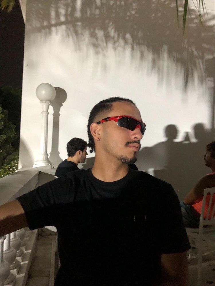 Homem pardo com camisa preta, olhando para o lado e mostrando o perfil do rosto, óculos vermelho, com cavanhaque e cabelo trançado