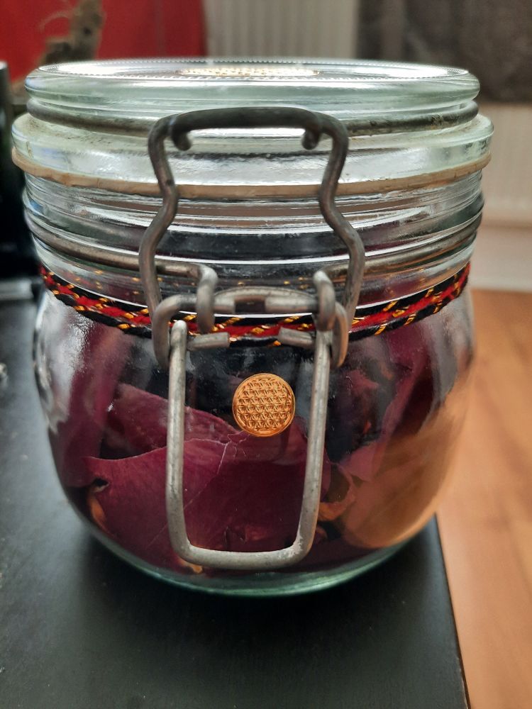 a closed glass jar full of dried petals with red and black thread around it and another smaller gold flower of life sticker on the front 