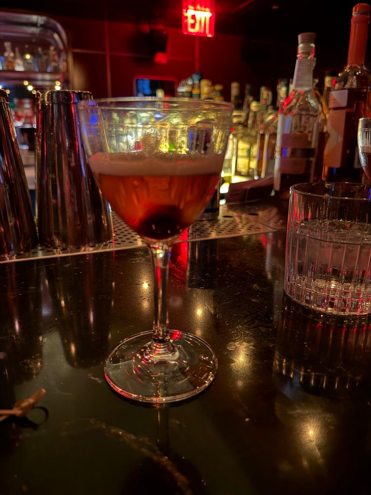 Manhattan drink in a small tulip glass on a bar in front of liquor bottles. 