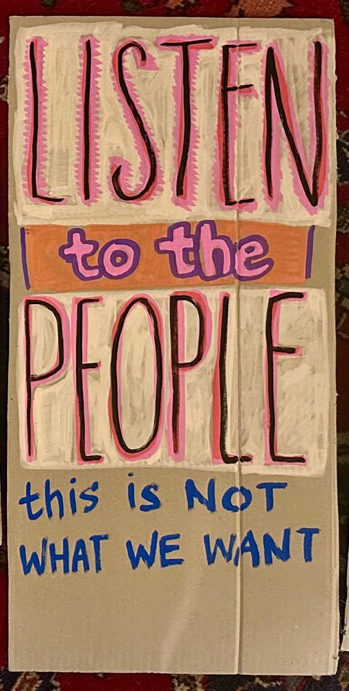 Text painted on a cardboard sign reading “LISTEN to the PEOPLE, this is not what we want”