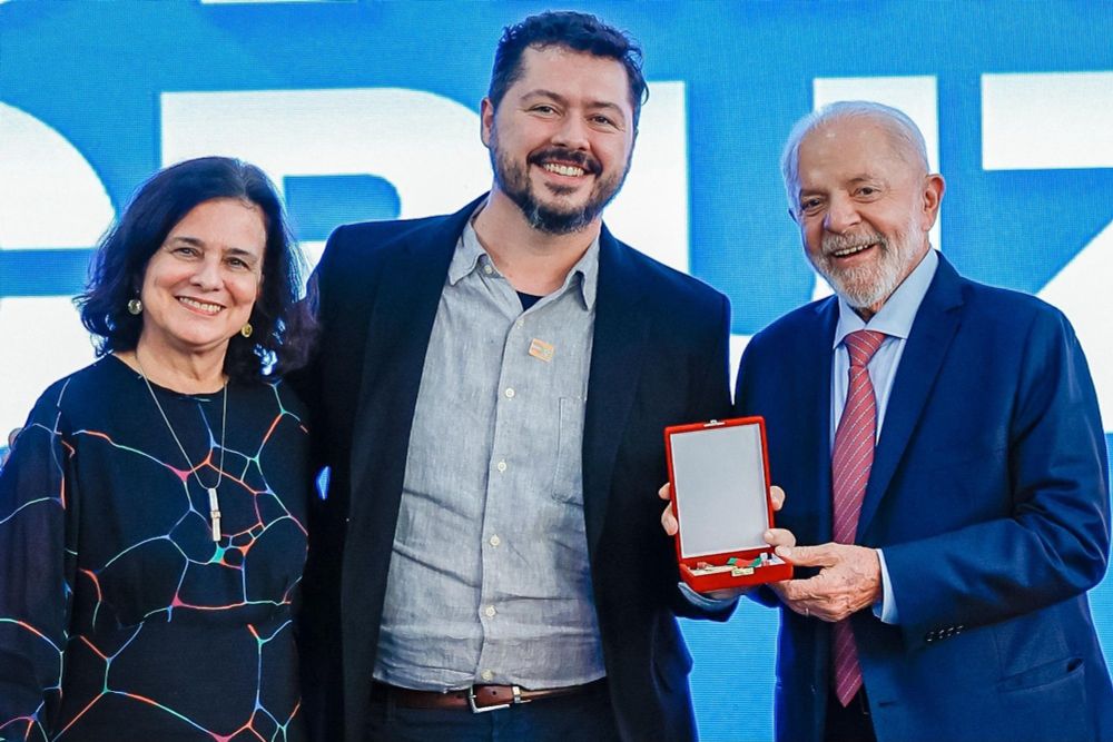Uma foto minha no evento ao lado da Ministra da Saúde Nísia Trindade e do Presidente Lula, que está segurando a medalha Oswaldo Cruz (uma caixa vermelha com uma medalha dourada pendurada por uma faixa verde e vermelha). Estou de paletó preto e camisa cinza entre os dois, enquanto encolho a barriga.