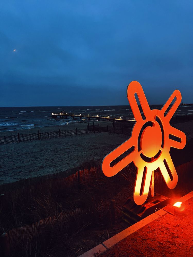 Foto zeigt eine angeleuchtete künstlerische Sonne in der Abendämmerung . Die Sonne hat sechs Strahlen. Im Hintergrund eine beleuchtete Seebrücke und die Ostsee. 