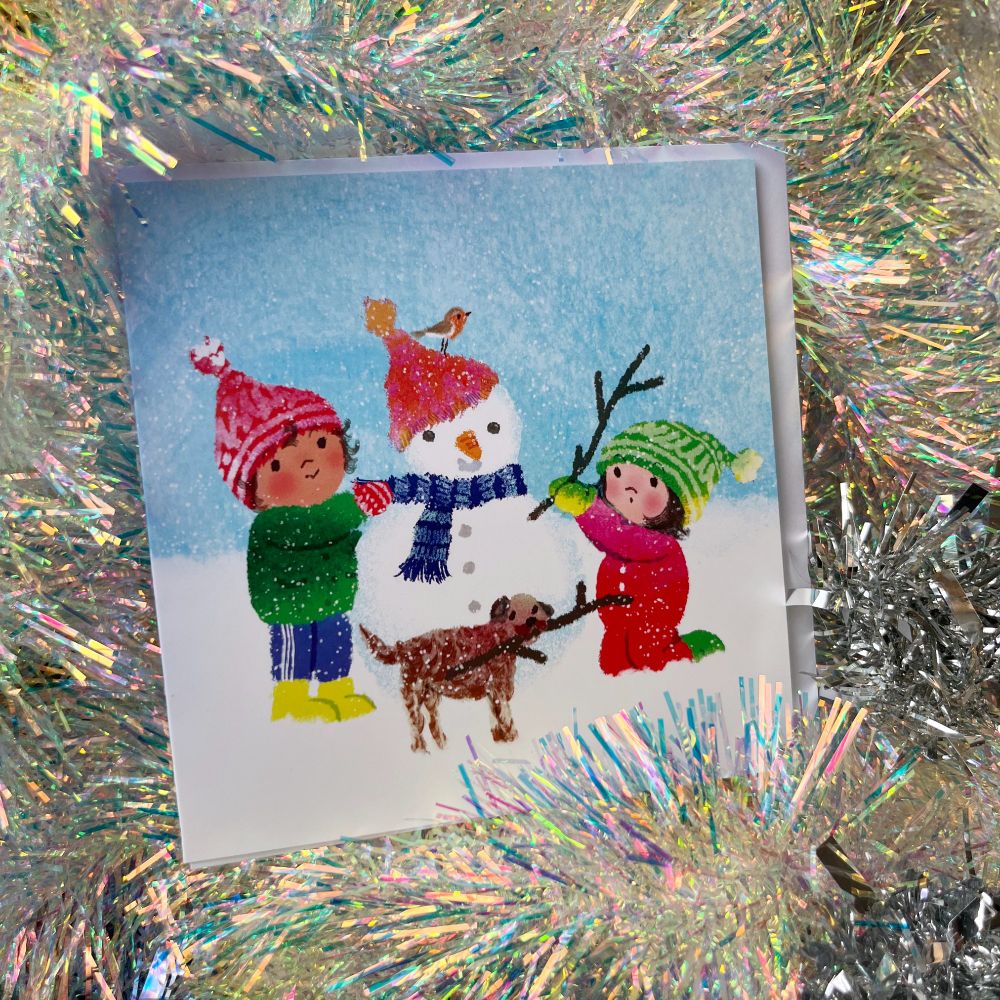 a photo of a christmas card by artist kip alizadeh showing two children wrapped up cosily building a snowman with their dog. on the snowman's hat a robin is perched.