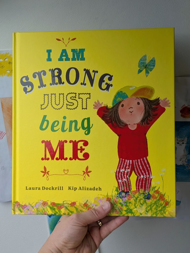 A photo of Kip's hand holding an advance copy of I Am Strong Just Being Me by Laura Dockrill and Kip Alizadeh. 