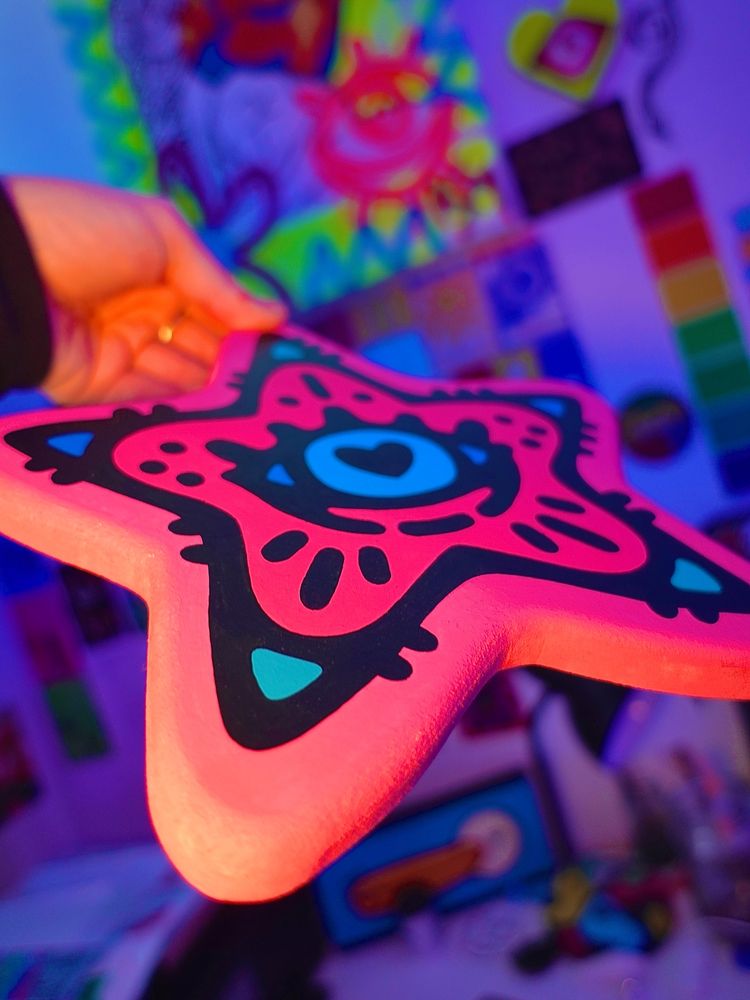 A picture of my hand holding a painted pink woodcut star shaped piece with a bold black star outline and an eye in the centre. The camera angle is from one of the points of the star