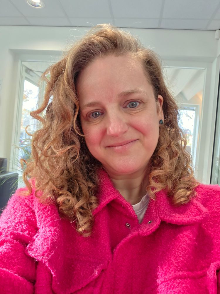 Een blonde vrouw met krullen heeft een felroze jas aan. De achtergrond is wit met een glazen deur half zichtbaar achter de vrouw. 