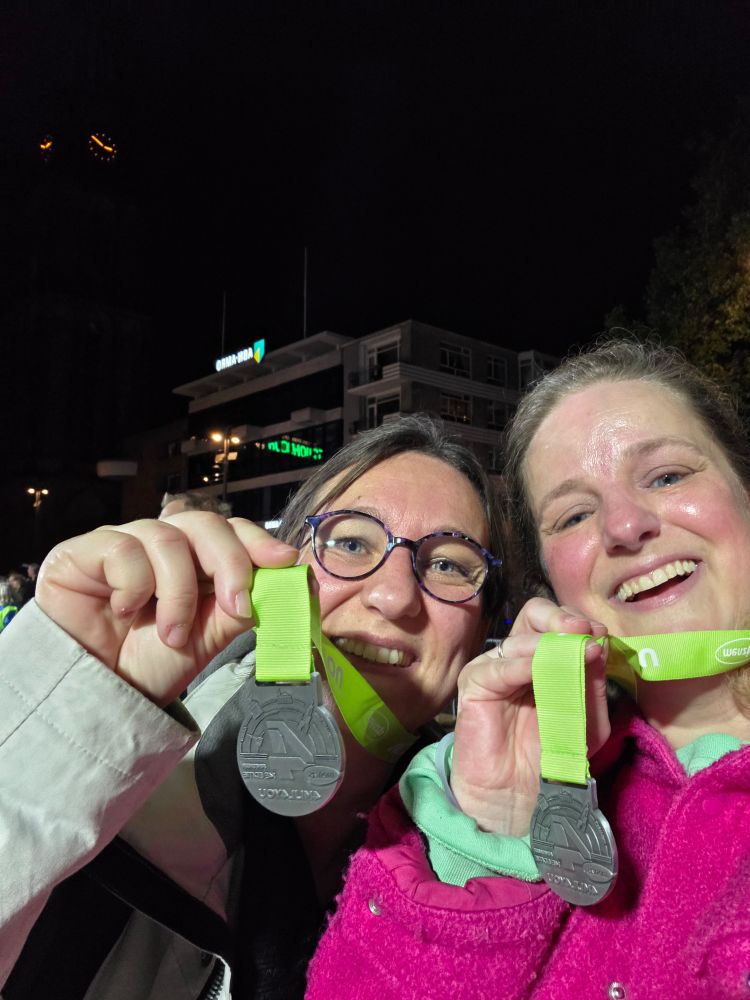 Twee vrouwen houden een medaille aan een gifgroen lint omhoog voor hun gezicht. Ze hebben rode wangen van de inspanning en de warmte. De vrouw rechts heeft een felroze jas aan waaruit een zeegroene mouw piept. De vrouw links heeft een donkere bril op en een grijze jas aan. Ze kijken allebei erg blij. De achtergrond is donker, maar je ziet nog vaag een kantoorgebouw en wat licht weerspiegelt in de ramen. 