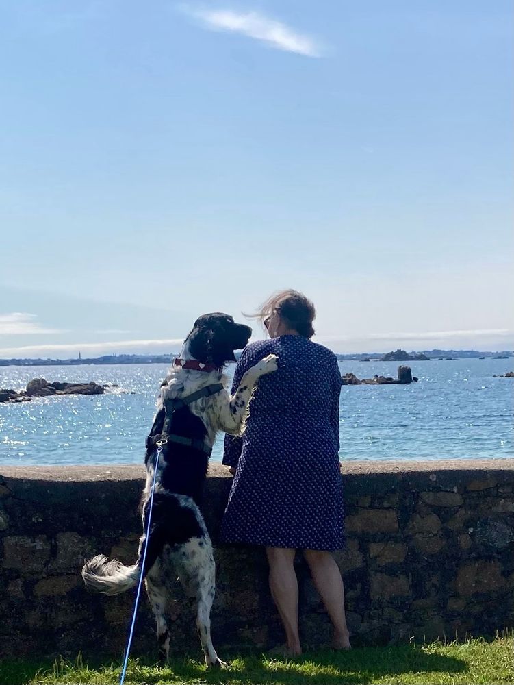 Hund und Frau stehen nebeneinander an einer Mauer und blicken aufs Meer. Der Hund hat eine Pfote auf die Schulter der Frau gelegt.