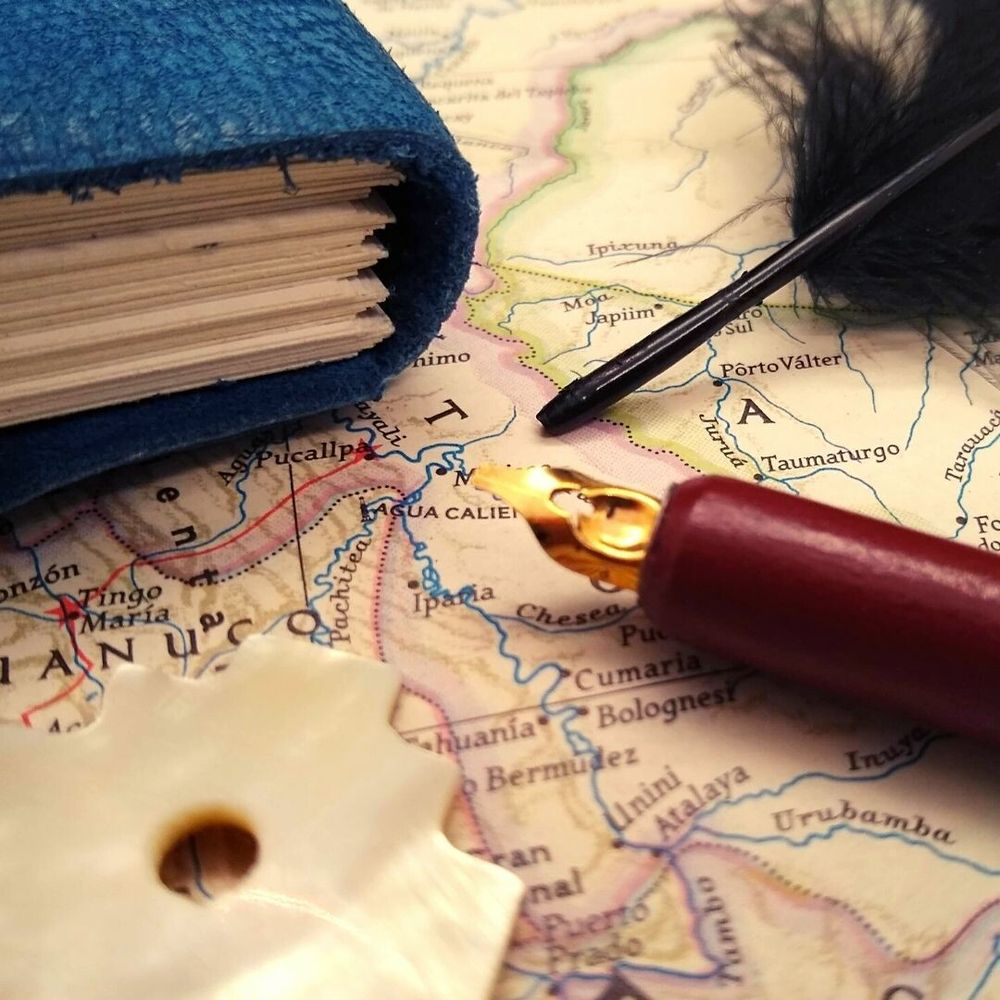 Eine historische Karte von Peru, auf der ein blaues Notizbuch, eine schwarze Feder, ein Kalligraphiefüller und ein perlmuttschimmernder Anhänger in Form eines Andenkreuzes liegen.