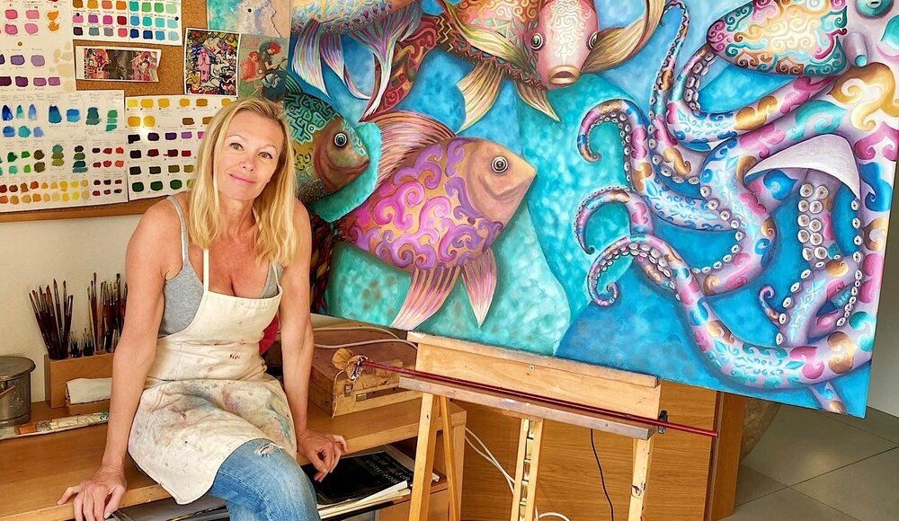 Alicia Zemanek, an exceptionally skilled artist, has created a remarkable piece titled "It is Me" This artwork beautifully encapsulates the essence of Key West and the Florida Keys, with its vibrant colors and intricate details that truly reflect the charm and appeal of the area.
The image shows a woman standing in front of a large painting.  She is holding a paintbrush in her right hand and is looking at the painting.  She is wearing a white apron over a grey tank top and blue jeans. The painting is of an octopus with its tentacles extended. The octopus is blue, purple, and gold and is swimming in a turquoise blue sea.  There are three fish in the foreground of the painting.  They are all different colors and have intricate patterns on their scales.  The woman's work space is visible behind her, with paint swatches and brushes. The painting is on an easel and there is a wooden table in front of her.  The woman looks proud of her work. 