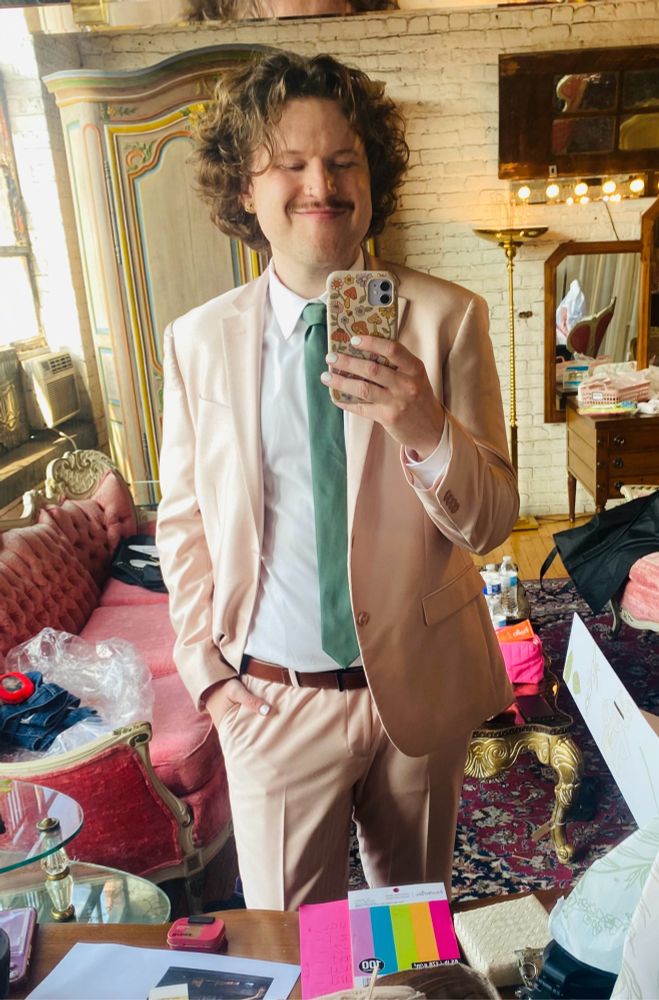 tuck selfie in a pink suit and green tie. She’s standing in a cluttered bridal suite