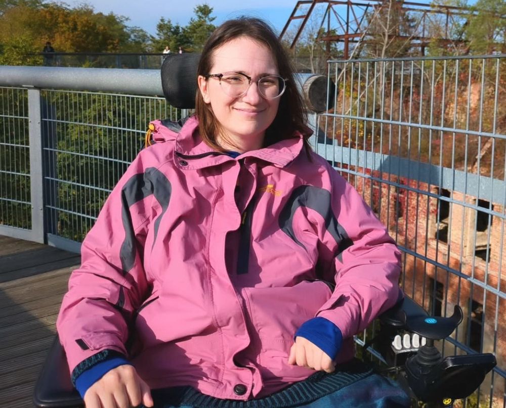 Bild von Tabea, die in die Kamera lächelt. Das Bild ist draußen aufgenommen. Die Sonne scheint ihr ins Gesicht. Tabea sitzt in einem E-Rollstuhl, trägt eine rosane Jacke, eine Brille und offene Haare.