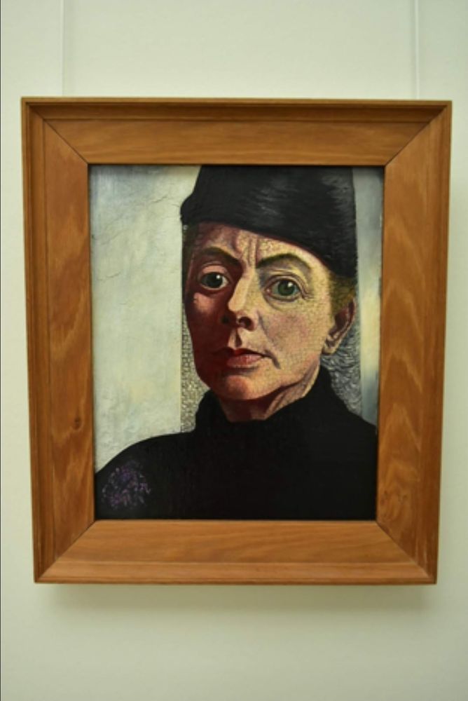 Zelfportret van Charley Toorop, een blanke vrouw in de rouw met een zwarte col en zwart randloos hoedje op. De grote blauwgroene ogen kijken je aan. De ene helft van het gezicht is belicht, ze heeft een scherpe neus en wat verticale rimpels boven de neus. De linkerkant is behalve rondom t oog, donker