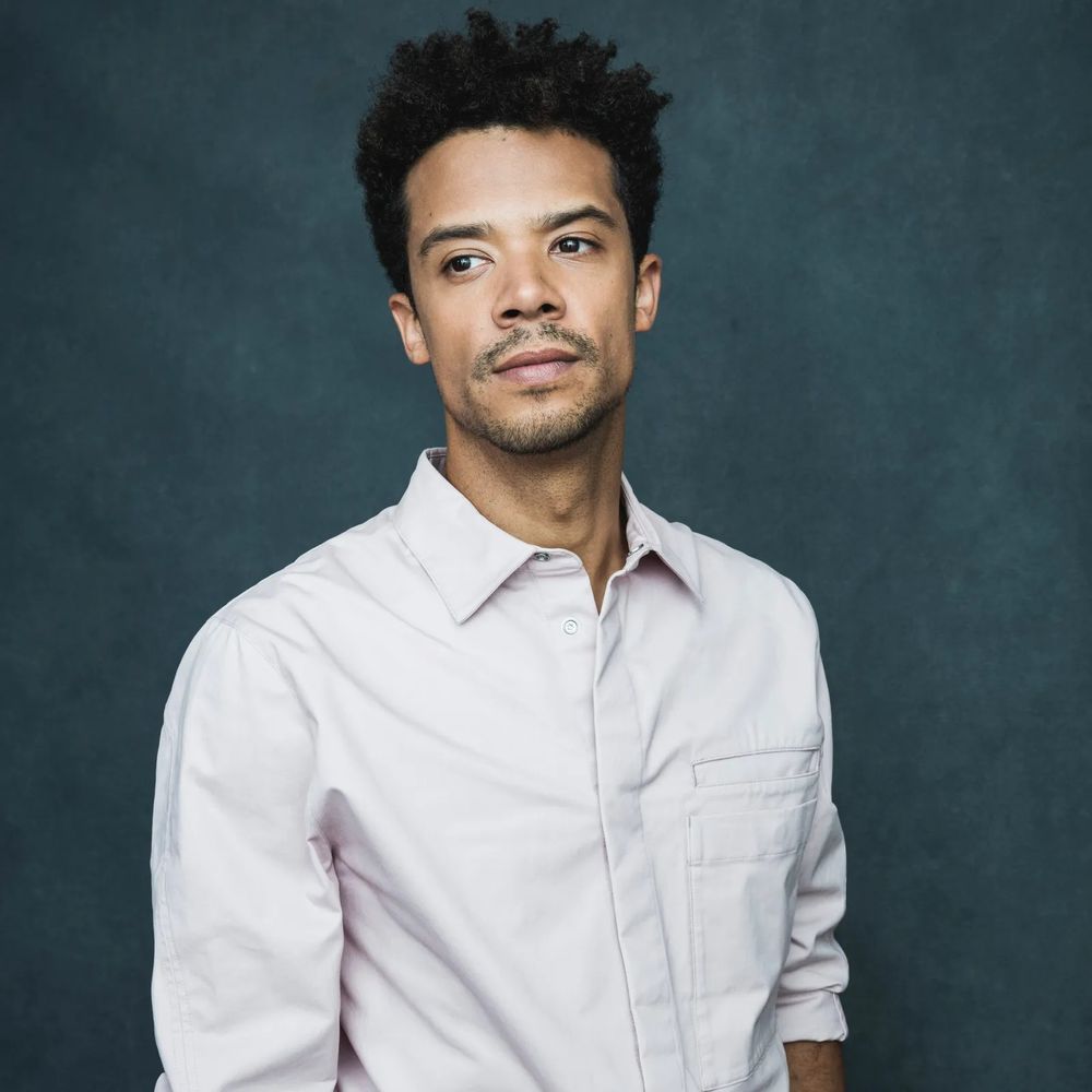Jacob Anderson in a button down shirt, looking off to the side with a serious look on his face.