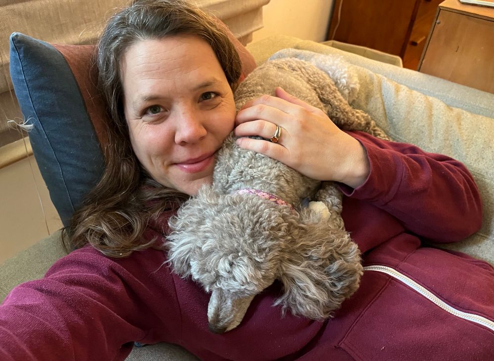 Brown-haired woman laying on a couch with. Brown mini poodle snuggled on her chest.