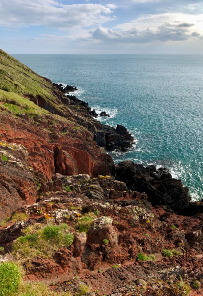 Coastal landscape at Manorbier Tenby, a rugged, rocky coastline with a dramatic cliff face rising up from the turquoise-colored waters of the ocean below. The cliffs have a mixture of reddish-brown and green hues, with patches of vegetation growing on the slopes. The sky is dotted with fluffy billowy "cotton-like" clouds