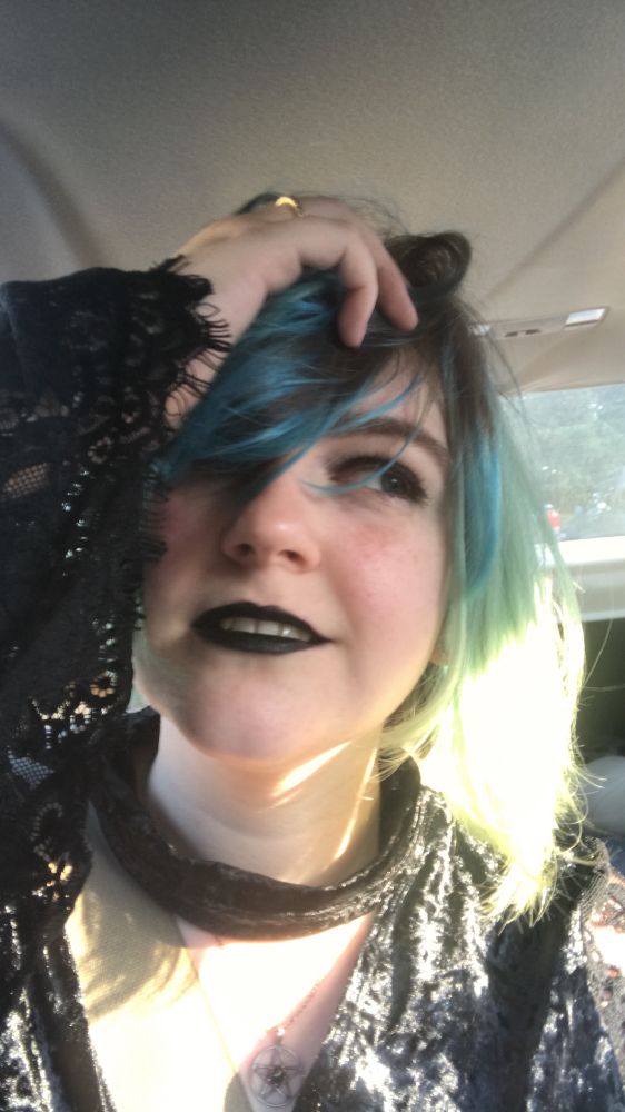 A woman with dark blue hair and dark makeup looks to the side