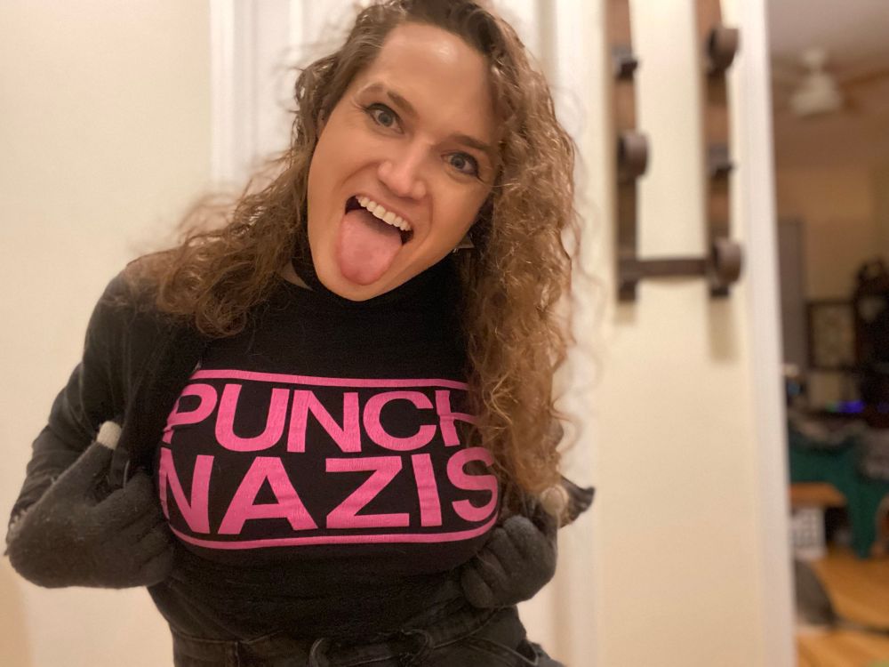 A woman pulls open her black jumpsuit to show the text of her shirt. In bright pink text a a black background it reads “PUNCH NAZIS.” 