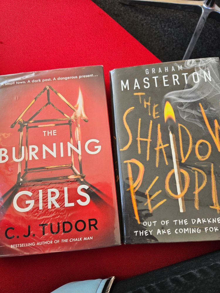 A photo of two books sitting on a red seat. The left is a red book titled The Burning Girls by C.J. Tudor while the second js a black book titled The Shadow People by Graham Masterton.