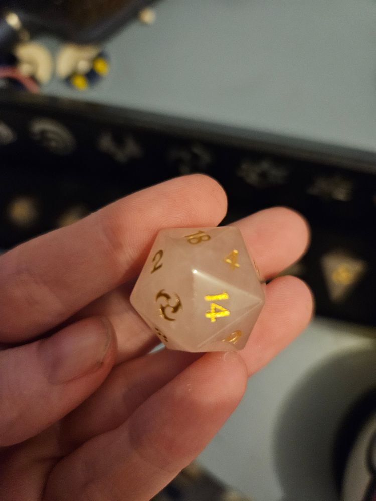 A pink dice made of Quartz in a person's hand. Its numbers are in gold with the "20" on the dice replaced with the symbol of Eletrco from Genshin Impact.