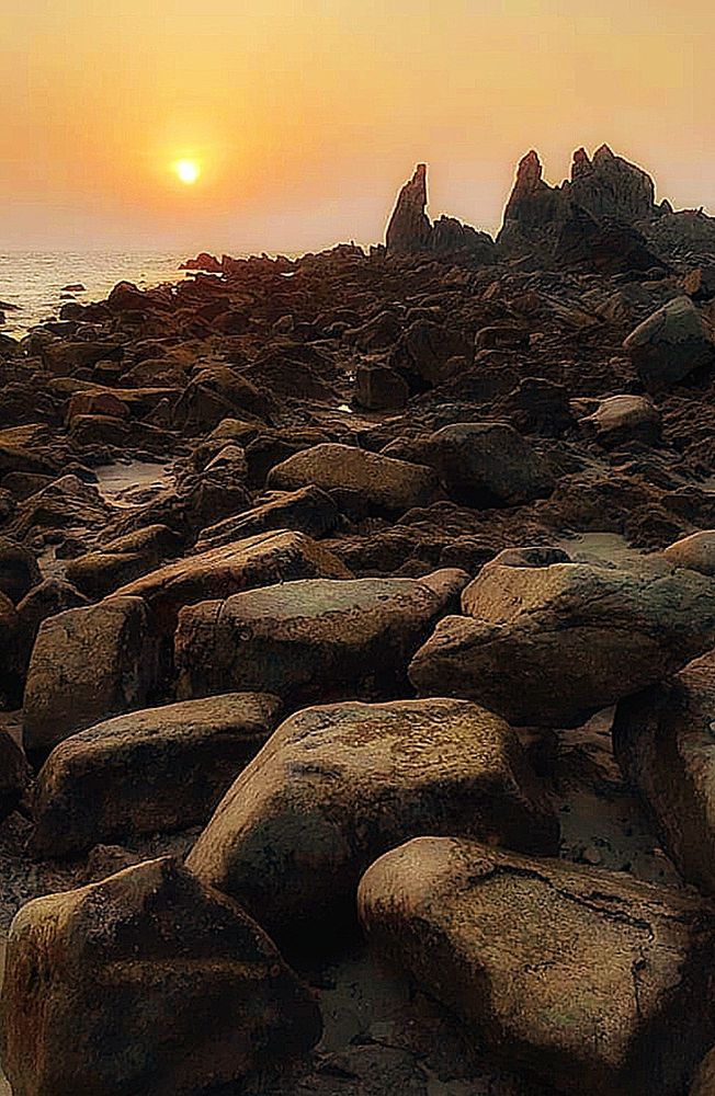 Ein Steinstrand bei Sonnenuntergang. Im Vordergrund große Steine, weiter hinten kleinere Steine und am Ende stehen große Steine ​​wie kleine Felsen. Lins oben im Bild ist ein klein wenig das Meer und im Hintergrund die untergehende Sonne, die den Himmel in orange färbt, zu sehen. 