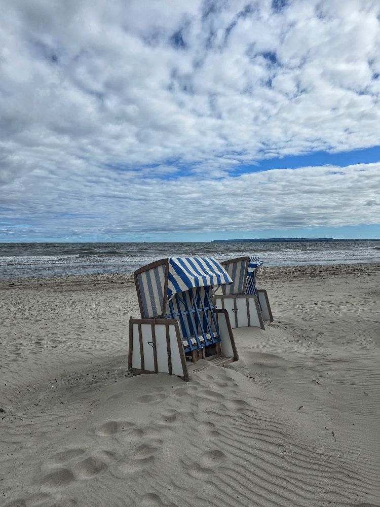 Two blue and white beach chairs stand on the Baltic Sea beach. Footprints are in the sand. Behind them is the Baltic Sea and a cloudy sky.