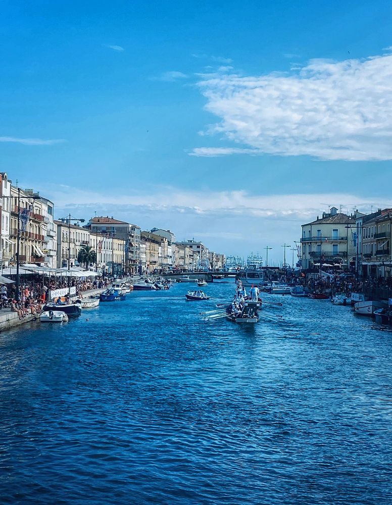 In the middle between the houses is a river with boats on it. The water and the sky are blue. The sky has a few clouds.