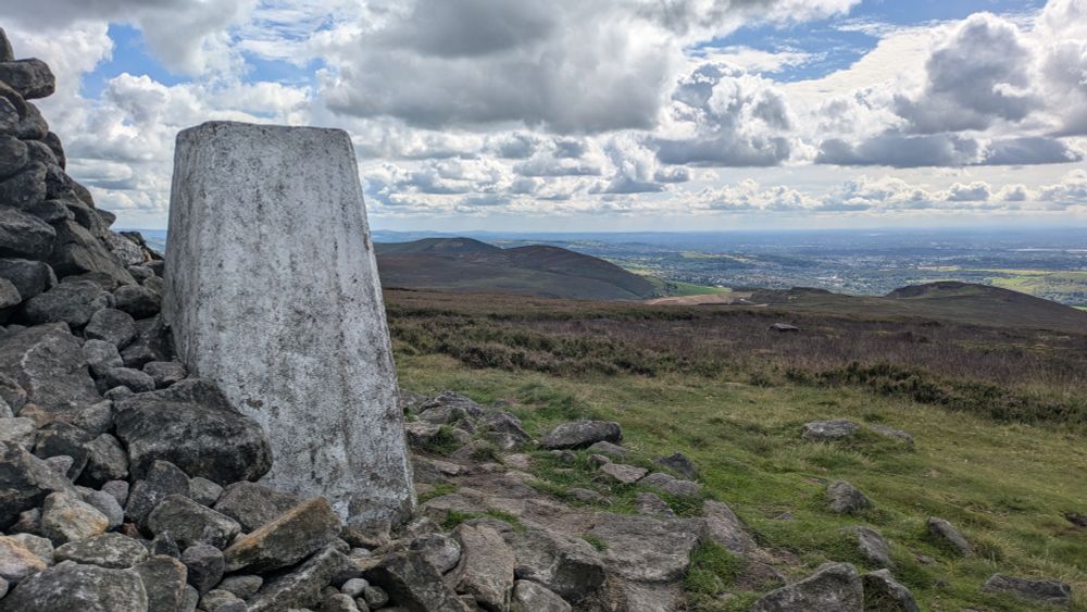 A trig point nestled against a pile of rocks. A clear view to the distant horizon. Fair weather cumulus clouds and blue sky.