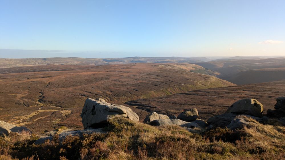 Rocky foreground looking down onto vast area of heather moorland. Sun shining from a cloudless blue sky.