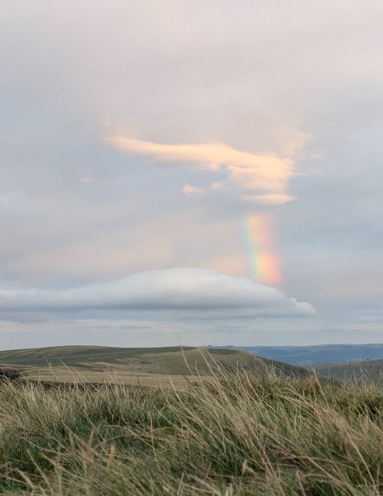Rough grass in the foreground. Sky is grey with a broken rainbow between two clouds.