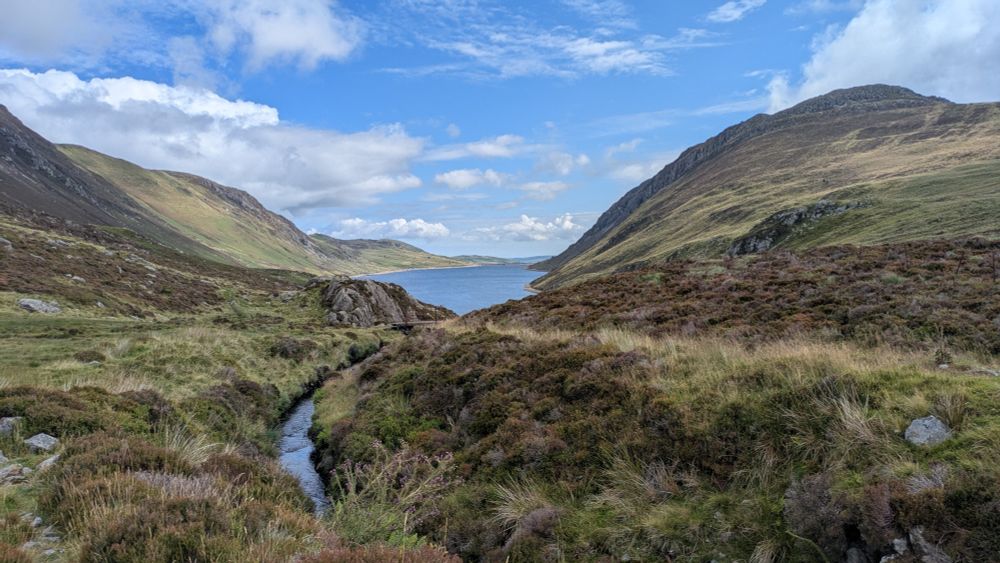 A stream leading to a reservoir between high hills. Blue sky with fair weather clouds.