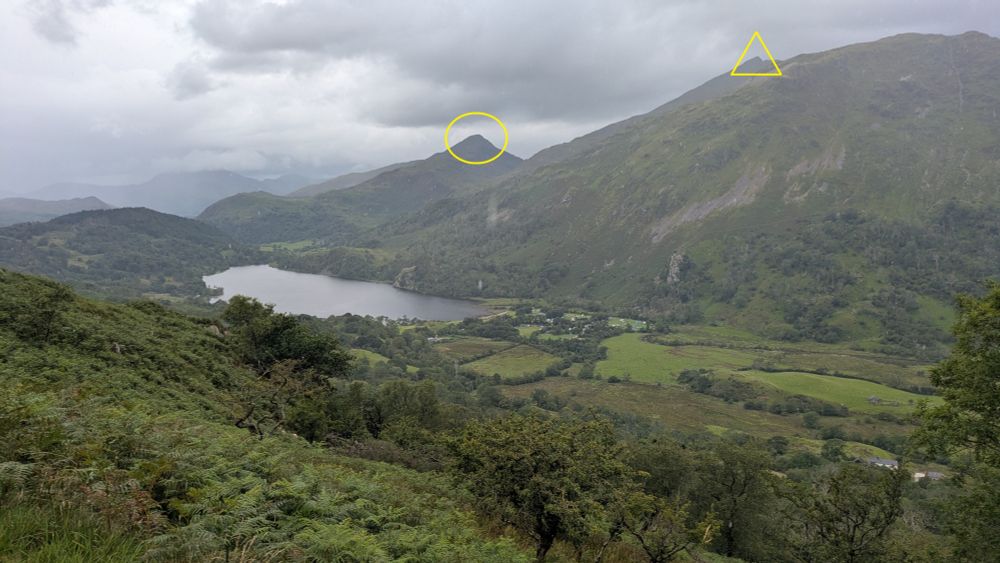 Looking down a steep bracken covered hillside to a flat grassy valley and lake. Tall mountains rise on the other side. The sky is grey and cloudy, threatening rain.