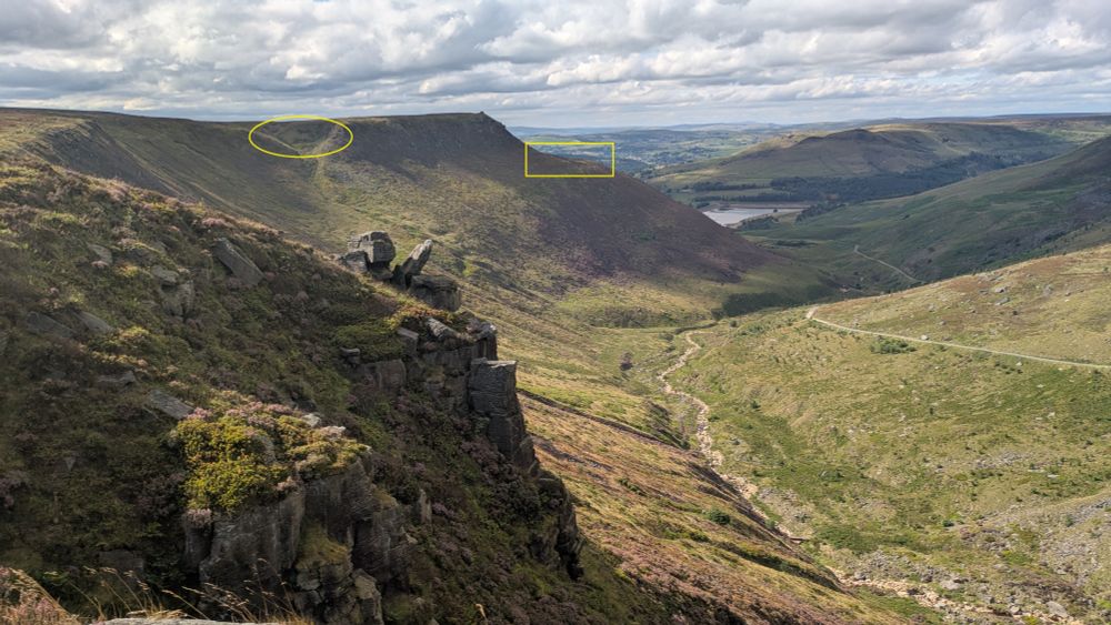 View from a hill looking down a valley to a distant reservoir. Very steep rocky ground immediately ahead with a stream and track in the valley. Partly cloudy.
2 features highlighted in yellow for map reading task - see accompanying map.