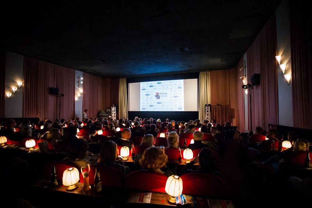 Der klassiche Kinosaal im Elite Kino Espelkamp mit hellroten Textilwandbespannungen und schwarzer Decke. Es ist dunkel, an den Wänden leuchten kleine dreieckige Lampen und auf den Tischchen vor den roten Kinosesseln kleine Schirmlämpchen.  Das Publikum schaut auf die von beigen Vorhängen gesäumte Leinwand, auf das ein spitziale-Logo projiziert wird.