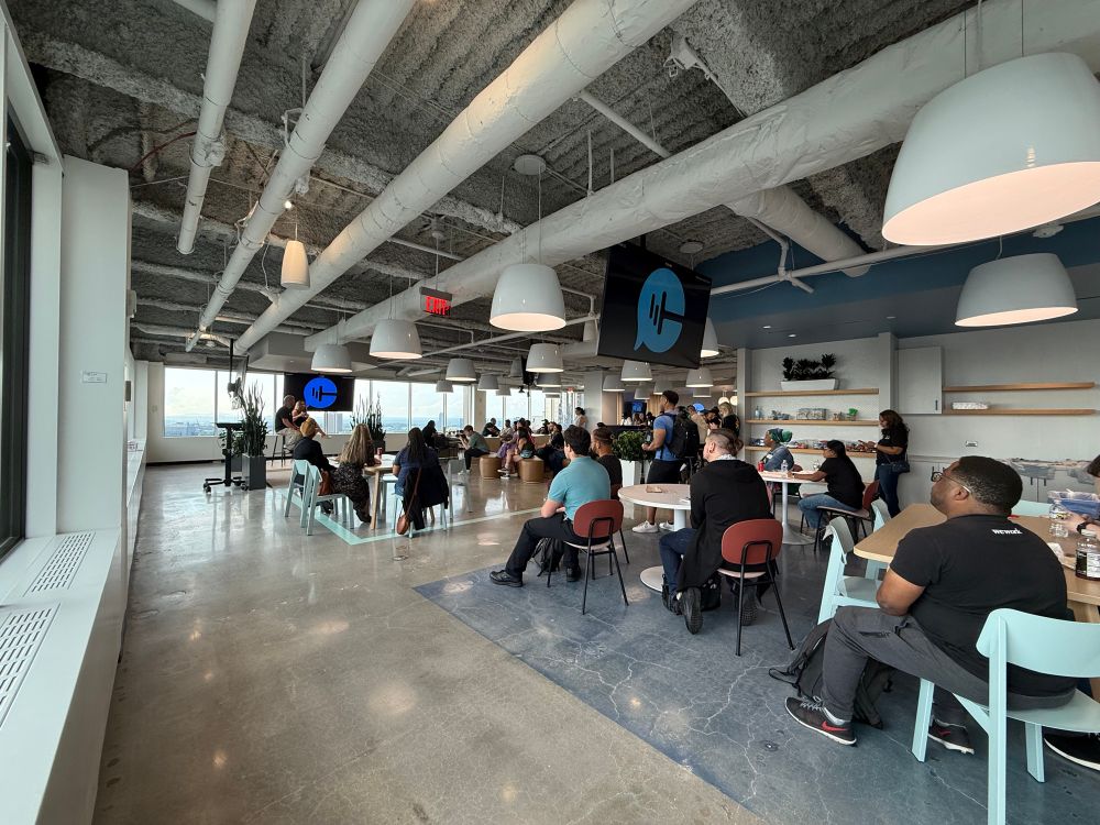 A digital screen mounted on a wall displaying the text “Meet Atlanta Tech Week & RenderATL Speakers!” with logos for ReactATL, Modern Web, Refactr.Tech, ATL, and JavaScript beneath the announcement. The background of the screen is a sleek mix of purples and blues with abstract shapes.