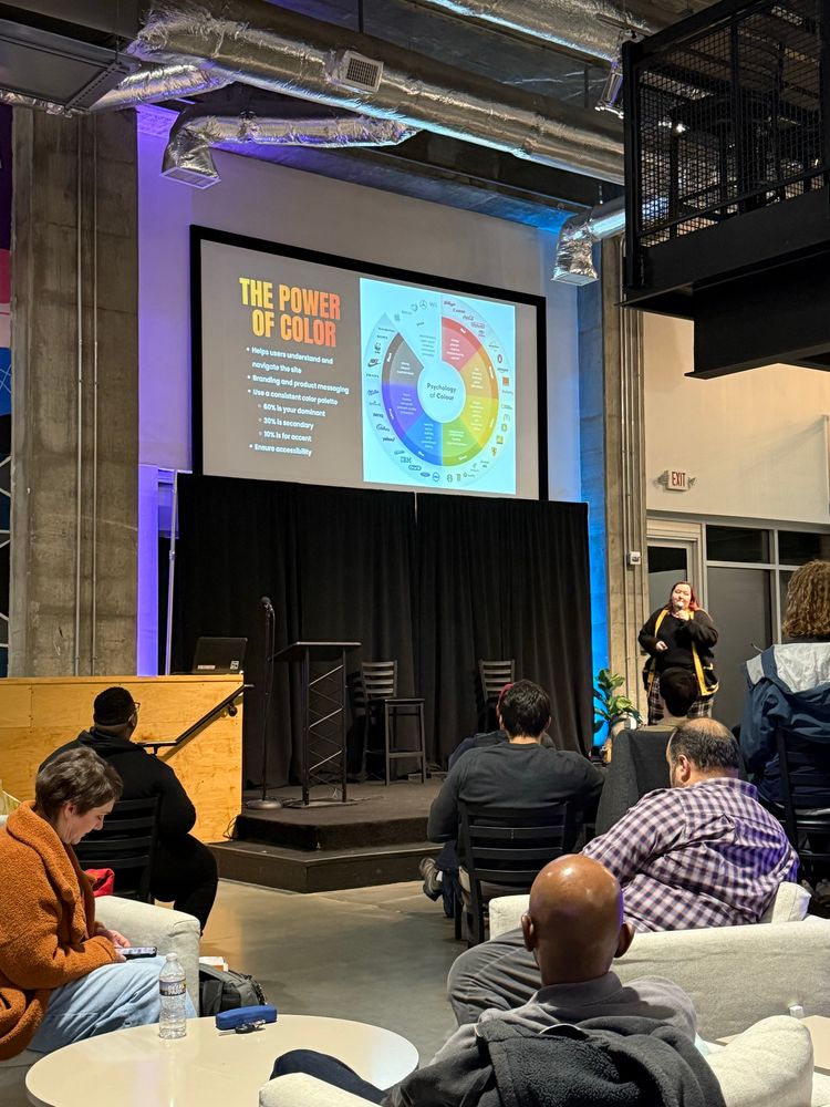 An indoor event with a presenter speaking on stage in front of an audience. The slide projected on the screen reads ‘The Power of Color’ with a color psychology chart displayed. The venue has industrial-style decor with exposed ducts and a casual seating arrangement. Attendees are seated, some looking at the speaker, while others are engaged with their devices.