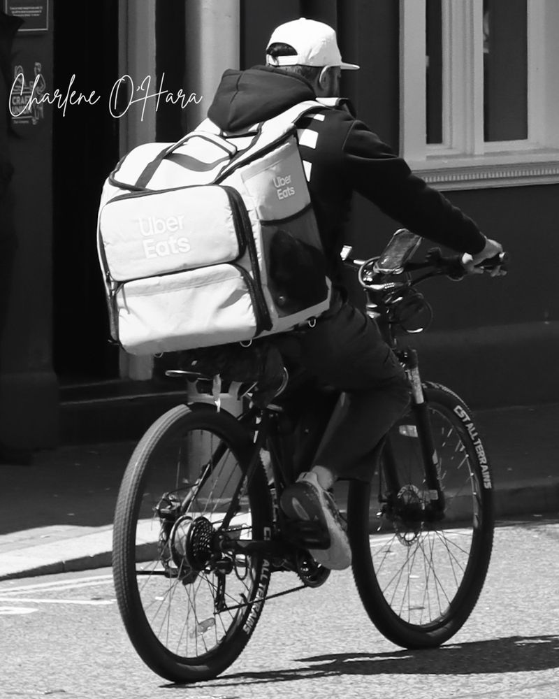 A black and white photo of a man on a bike. The man is wearing a baseball cap, a black hoodie with black trousers and white trainers. On his back is a backpack with the Uber Eats logo