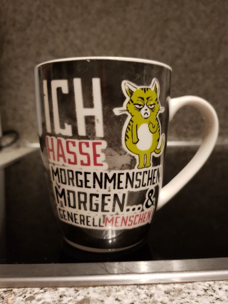 Tasse, bedrückt mit der Aufschrift "Uch hasse Morgenmenschen, Morgen & generell Menschen ". Daneben eine grüne Comic-Katze im Grinch-Style, die den Stinkefinger zeigt.