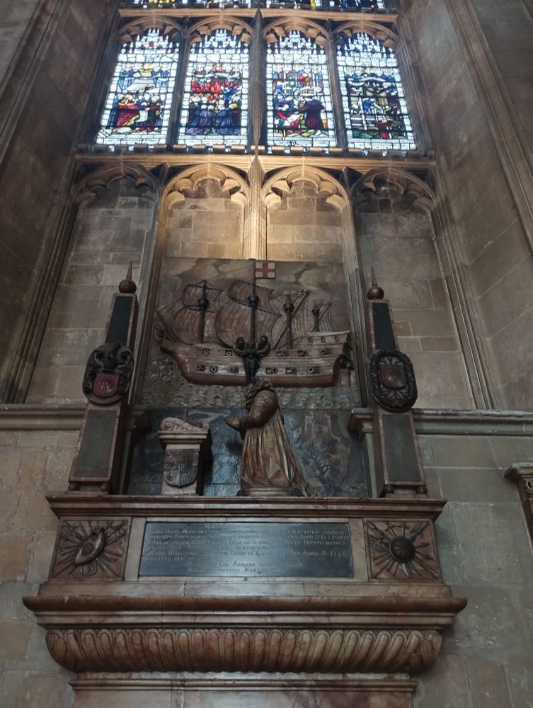 A carving of a ship with a carving of a lady kneeling in front, some stained glass windows above 