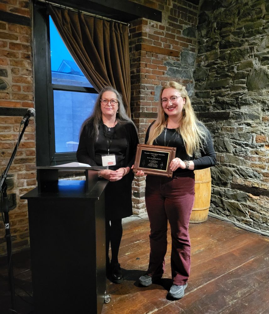 Two women, both namer Katie, are standing beside a podium. The older woman on the right has long grey hair and is wearing a black dress. The younger woman on the right has long blonde hair, a black long sleeved top, and burgundy pants, and is holding a plaque. They are in a brick and stone room with a wood floor. 