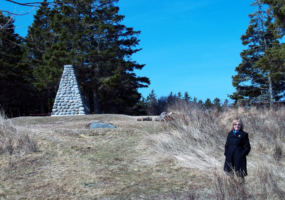 A woman with blonde cropped hair stands in a grassy field near a stone cairn monument, wearing a black overcoat. There are pine trees in the background and a blue sky. 