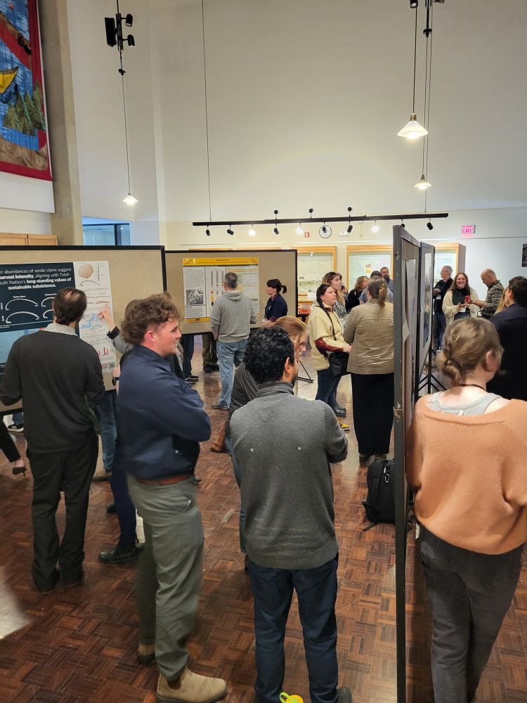 A group of people standing around in a large room, filled with rolling display boards. The boards have research posters on them, and the room is lit with warm lighting. This one is vertical, similar scene. 