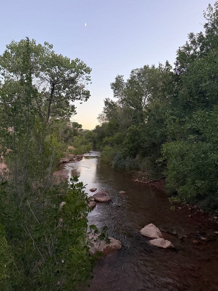 A dark river with rocks in the water and trees on both sides. The sky light blue to yellow fade with a crescent moon visible in the center. 