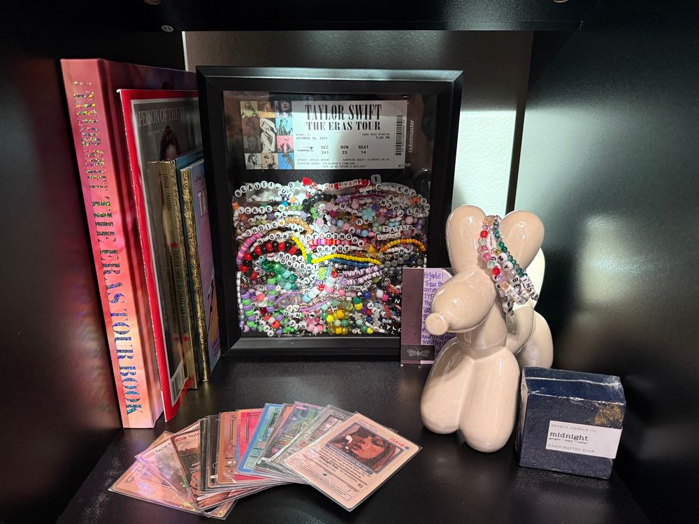A collection of Taylor Swift trading cards with her signature photo from midnights on the front, a dark blue soap with gold glitter and a label saying “midnight”, a pearlescent ceramic balloon animal with beaded friendship bracelets on it, a shadow box filled with beaded friendship bracelets and a replica concert ticket for the eras tour, with the eras tour book, time magazine, and two Taylor Swift little golden books. 