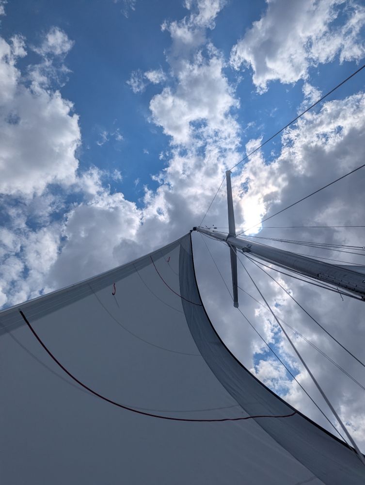 Looking up from the deck of a sailboat at the mast and sail with a blue sky and puffy white clouds in the background.