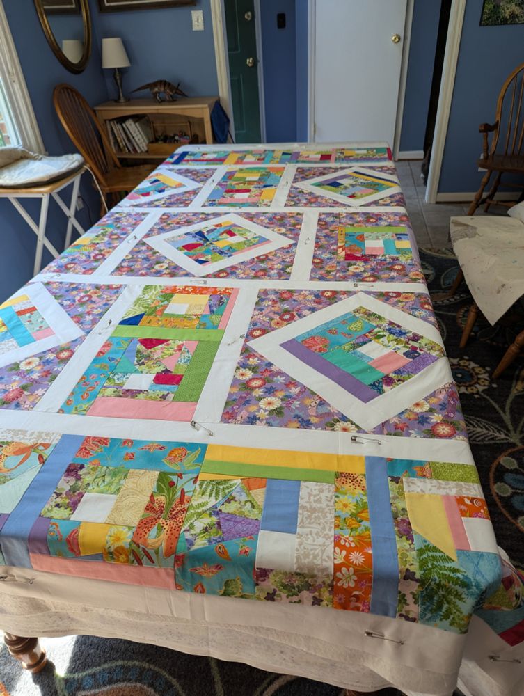 A quilt top of pastel crazy-quilt
 blocks and a lavender floral fabric with white sashing. It's laid out on a large dining table to be "sandwiched" and pin-basted with its batting and backing fabric, to get it ready for machine quilting. The background is a dining room with blue walls 