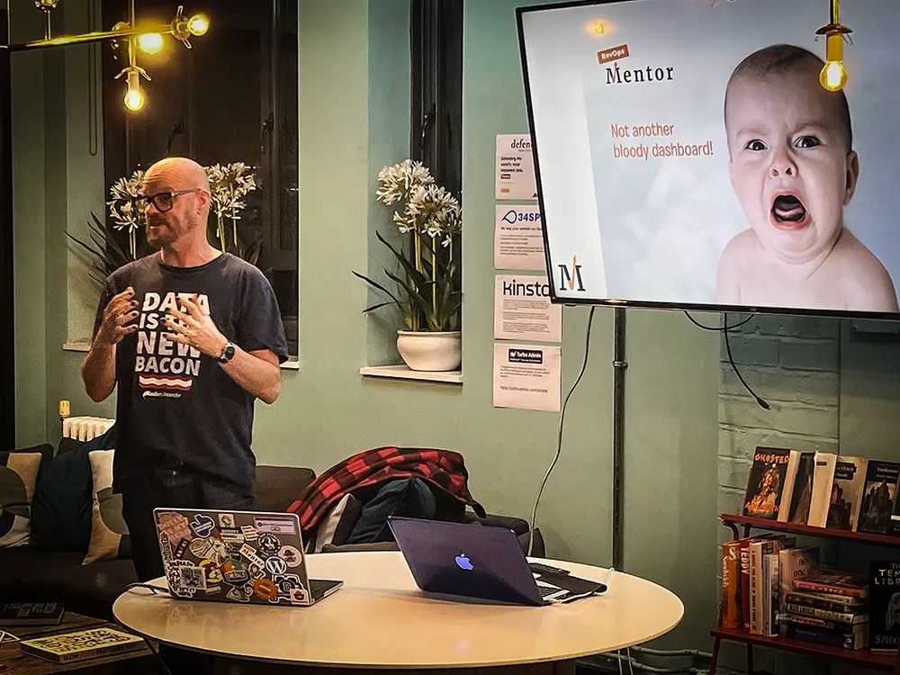 Jon Payne stands in a cozy room in front of a round table with two laptops, speaking to an audience. he is wearing a shirt that reads "DATA IS THE NEW BACON." Behind him, a presentation slide is displayed on a large screen with the heading "Mentor" and the phrase "Not another bloody dashboard!" partially visible next to an image of a baby.