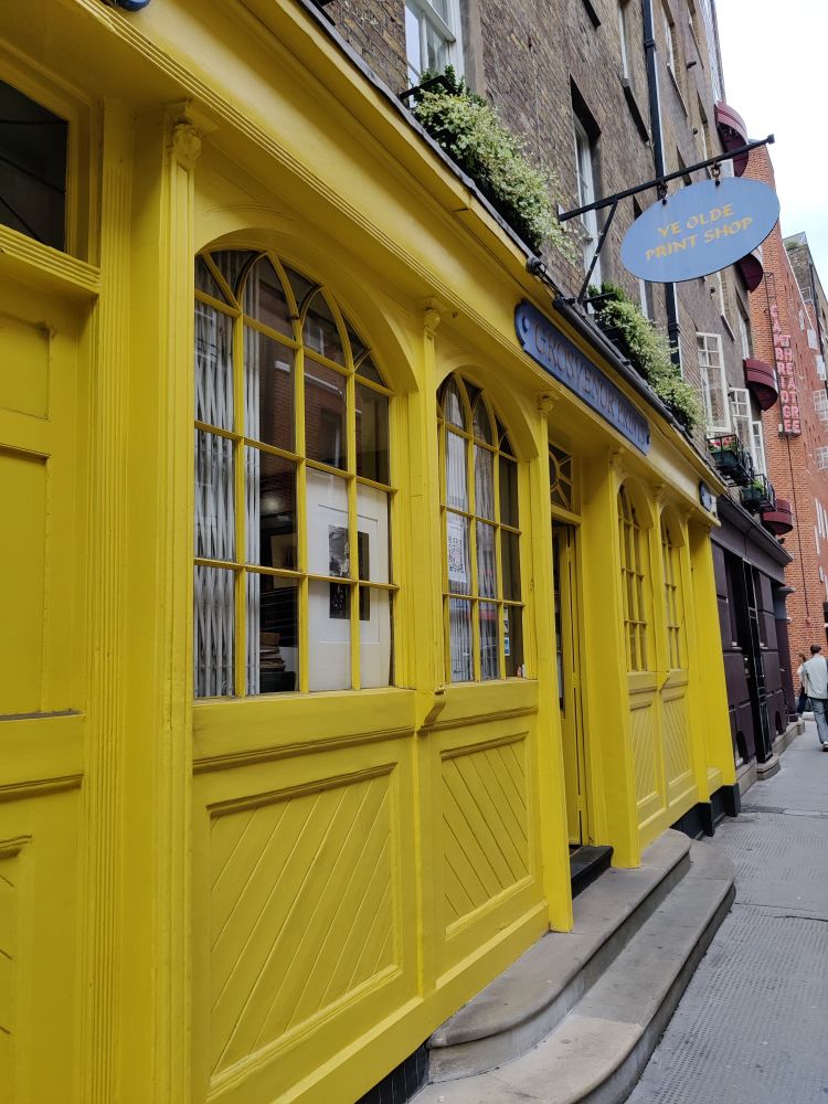 A small old fashioned looking wooden shop front painted bright yellow with a hanging sign that reads: Ye Olde Print Shop