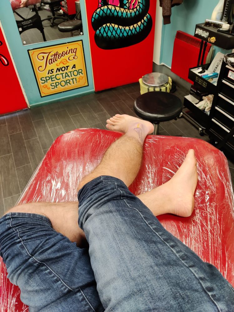 My bare lower legs laid out on a plastic clad red bed in a tattoo parlour. A faint stencil of a wing is visible on the outside of the right ankle. A sign is visible in the background "Tattooing is not a spectator sport"